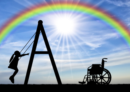 Silhouette of a child disabled girl on a swing next to a wheelchair on a background of the sky with a rainbow. Concept of playing children with disabilitiesの写真素材