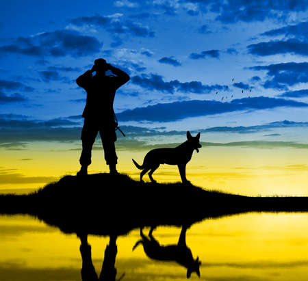 A Ukrainian military man with a dog looks through binoculars against the background of the sky in the form of the Ukrainian flag - a silhouette. Concept of Ukraine's war with Russiaの写真素材