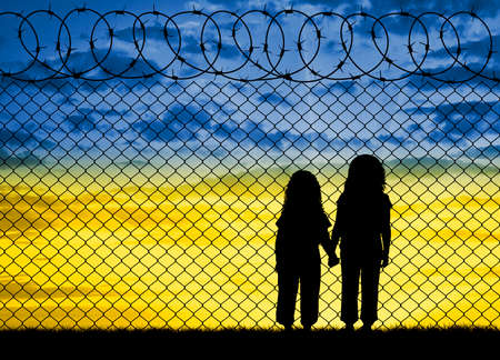 Ukrainian refugees - children, at the barbed wire fence against the sky in the form of the flag of Ukraine. Concept of Ukrainian refugees. Crisis and war in Ukraine with Russiaの写真素材