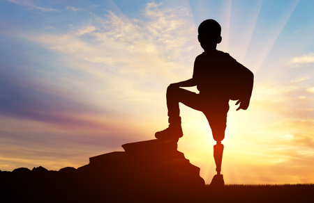 Silhouette of a disabled child with a prosthetic leg standing triumphantly on a rock against a stunning sunset backdrop. Perfect for themes of overcoming adversity, motivation, and inspiration.の写真素材