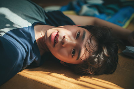 A young Asian boy is lying on a wooden floor in the sunlight, looking up weakly, possibly feeling dizzy or having fainted from a child heart problem.の素材