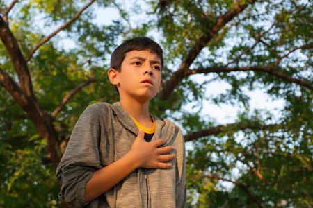 A young boy stands under a large tree, looking up with a worried expression and his hand over his heart, suggesting a symptom of a child heart disease.の素材