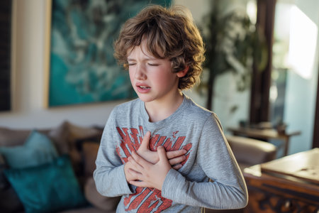 A young boy with curly hair and eyes shut tight clutches his chest with both hands, suffering from an intense and painful symptom of a child heart disease.の素材