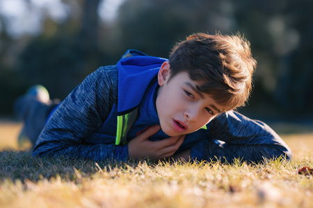 A young boy lies on the grass, looking weak and holding his chest after physical activity. This is a potential emergency symptom of a child heart disease.の素材