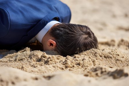 A businessman in a full suit has his head buried in the sand on a beach, a concept for ignoring problems, denial, or avoiding difficult situations.の素材