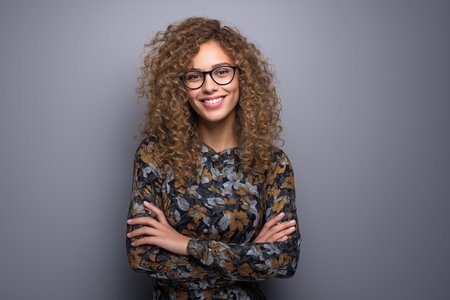 A studio portrait of a happy mixed-race woman with arms crossed against a gray background. Her genuine smile and stylish look express positivity and self-assurance.の素材