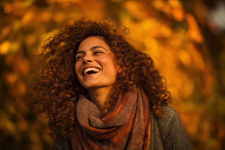 A happy mixed-race woman with a hearty laugh enjoys a beautiful genuine fall day. The golden, blurred background of autumn leaves enhances her joyful mood.の素材