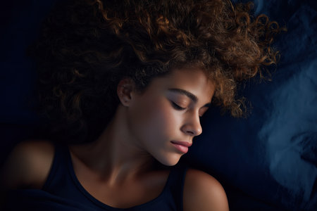 A serene close-up of a beautiful young mixed-race woman with voluminous curly hair, sleeping peacefully with her eyes closed on a dark blue pillow.の素材