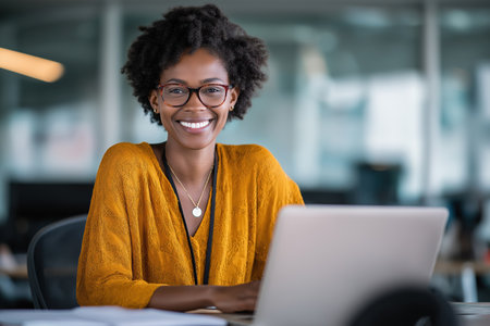 A happy and professional young black woman with an afro and glasses works on her laptop in a modern office, looking at the camera with a positive attitude.の素材