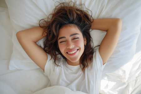 A beautiful and refreshed young woman lies on her back in a white bed, smiling with her eyes closed and hands behind her head after a good night's sleep.の素材
