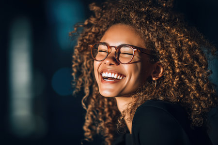 A beautiful mixed-race woman with amazing curly hair and glasses is laughing with pure joy, her eyes closed, against a dark background with city bokeh lights.の素材
