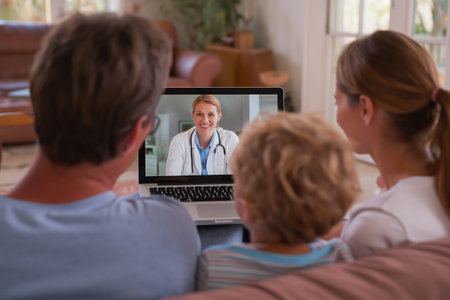 A mother, father, and their young child sit together on a sofa, having a group telehealth consultation with a smiling female doctor on their laptop screen.の素材