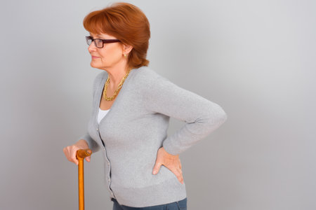 Mature woman in glasses holding a walking stick and grimacing due to sharp lower back or hip pain. Concept: aging, mobility, and chronic joint issues.の素材