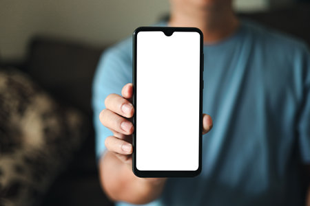 Smartphone blank screen presentation. A young man is holding a phone with a white empty screen mockup. a man is wear blue t-shirt in living room at home.の写真素材