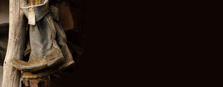 Old rustic boots hang on a pillar under the roof of the house. Vintage shoes. Dark background with...