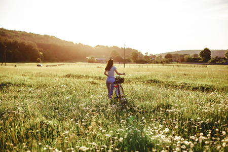 The girl walks with a puppy in a field in a bicycle in the back of warm, sunny lightの写真素材