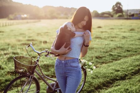 The girl walks with a puppy in a field in a bicycle in the back of warm, sunny lightの写真素材