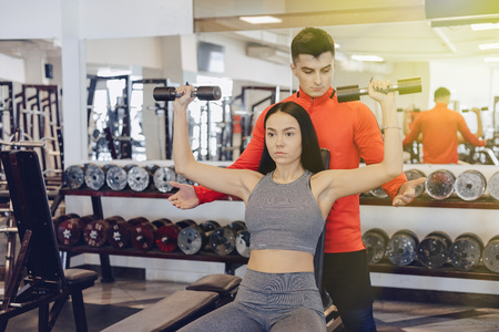 A young girl wearing sportswear in a gym performs dumbbell exercises, the coach helps her, sports, healthy lifestyleの写真素材