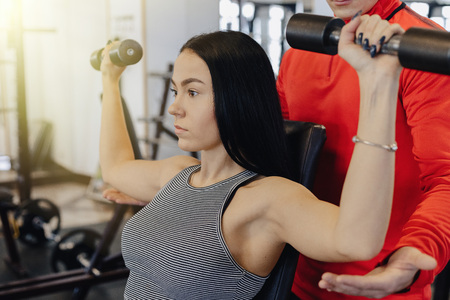 A young girl wearing sportswear in a gym performs dumbbell exercises, the coach helps her, sports,...