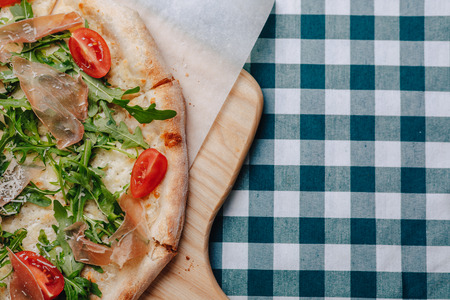 Neapolitan pizza on a cream sauce with ham, cheese, arugula, basil, tomatoes sprinkled with cheese on a wooden board on a tablecloth in a cell with a place for textの写真素材
