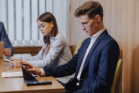 A stylish young man in a jacket and shirt sits at the desk with his colleagues and works with documents at officeの写真素材