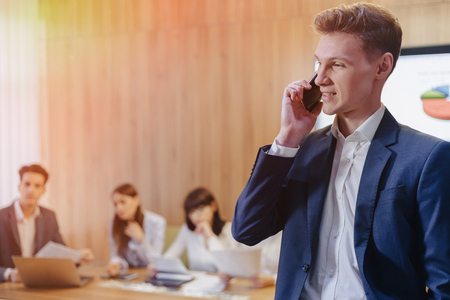 Stylish young businessman wearing a jacket and shirt on the blurry background of a working office with people talking on a mobile phoneの写真素材
