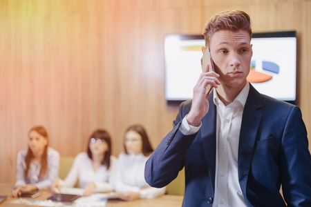 Stylish young businessman wearing a jacket and shirt on the blurry background of a working office with people talking on a mobile phoneの写真素材
