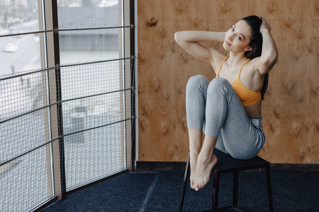 Young attractive fitness girl sitting on chair near the window on the background of a wooden wall, resting on yoga classes at gymの写真素材