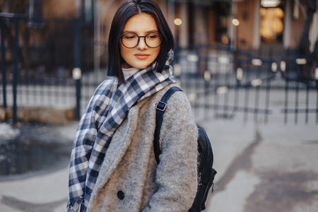 Attractive young girl wearing glasses in a coat on a urban background walking on a sunny dayの写真素材