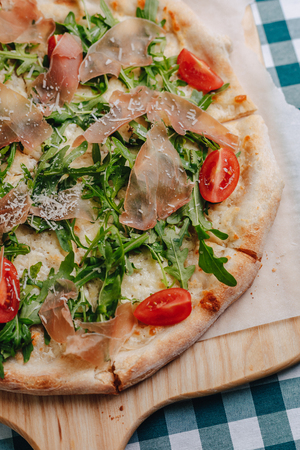 Neapolitan pizza on a cream sauce with salami, arugula, tomatoes sprinkled with cheese on a wooden board on a tablecloth in a cell with a place for textの写真素材