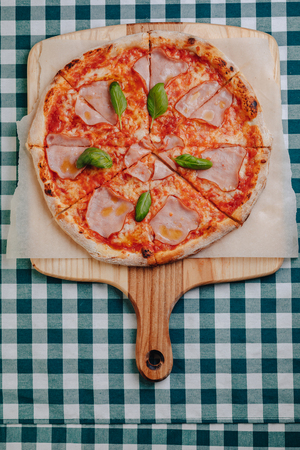 Neapolitan pizza on a cream sauce with ham, cheese, arugula, basil, tomatoes sprinkled with cheese on a wooden board on a tablecloth in a cell with a place for textの写真素材