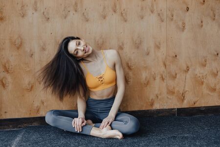 Young attractive fitness girl sitting on the floor near the window on the background of a wooden wall, resting on yoga classes at gymの写真素材