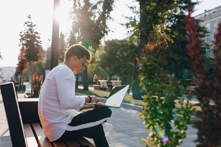young stylish guy in white shirt with phone and notebook works on bench on sunny warm day outdoors, freelanceの写真素材
