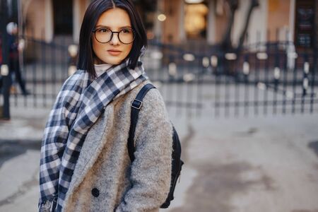 Attractive young girl wearing glasses in a coat on a urban background walking on a sunny dayの写真素材