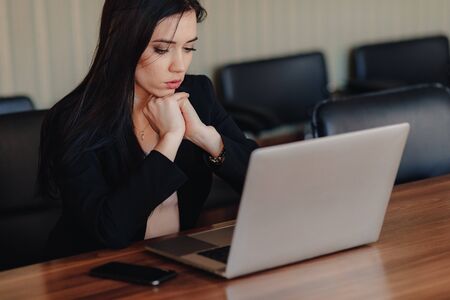 Young attractive emotional girl in business-style clothes sitting at a desk on a laptop and phone in the office or auditorium aloneの写真素材