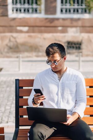 young stylish guy in white shirt with phone and notebook works on bench on sunny warm day outdoors, freelanceの写真素材