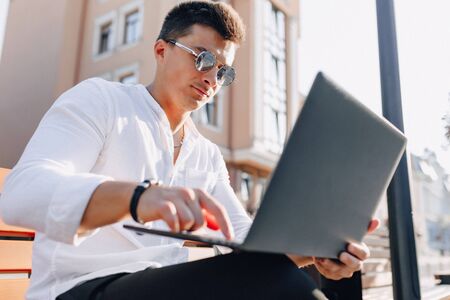 young stylish guy in white shirt with phone and notebook works on bench on sunny warm day outdoors,...