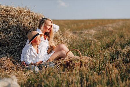 pretty blond long-haired woman with little blond son at sunset relaxing in the field and savoring fruit from a straw basket. summer, farming, nature and fresh air in the countryside. sunset.の写真素材