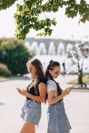 two young pretty girls on a walk in the park with phones. warm sunny summer day, joy and friendships.の写真素材