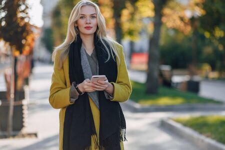 cute emotional attractive blonde woman in coat with smartphone walks the city street. communication during the walk, lifestyle, street in good weather.の写真素材