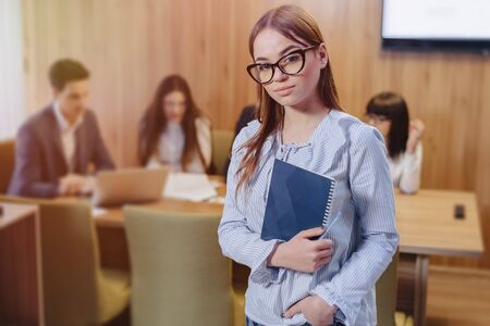 young attractive stylish office worker girl in glasses with a notebook in the hands on blurry background of working colleaguesの写真素材