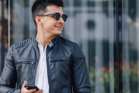 young stylish guy in glasses in black leather jacket with phone on simple glass backgroundの写真素材