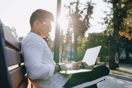 young stylish guy in white shirt with phone and notebook works on bench on sunny warm day outdoors, freelanceの写真素材