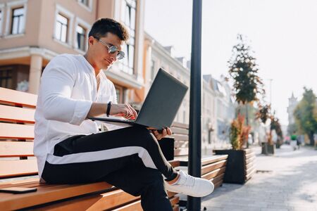 young stylish guy in white shirt with phone and notebook works on bench on sunny warm day outdoors, freelanceの写真素材
