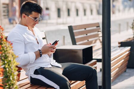 young stylish guy in white shirt with phone on bench on sunny warm day outdoorsの写真素材