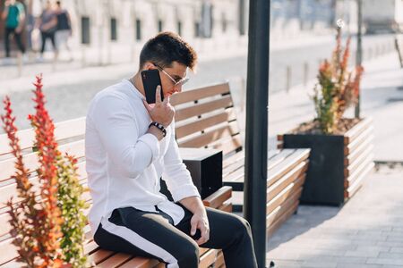 young stylish guy in white shirt with phone on bench on sunny warm day outdoorsの写真素材
