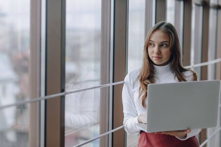 pretty young girl with laptop by the window in office with big windows. remote work concept....