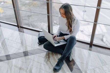 attractive girl working with laptop and things in airport terminal or office on floor. travel atmosphere or alternative work atmosphere. freelancer student travels to business meeting. concept of alternative workplace, technology and waiting.の写真素材