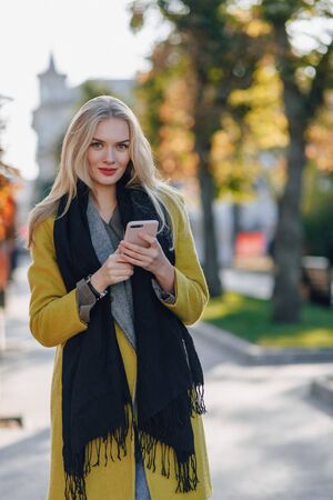 cute emotional attractive blonde woman in coat with smartphone walks the city street. communication during the walk, lifestyle, street in good weather.の写真素材
