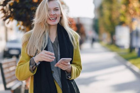 cute emotional attractive blonde woman in coat with smartphone walks the city street. communication during the walk, lifestyle, street in good weather.の写真素材